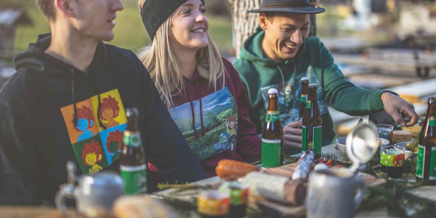 Lachende Freunde genießen eine bayerische Biergarten-Stimmung mit rustikalem Esstisch, bedeckt mit Speisen und Bier.