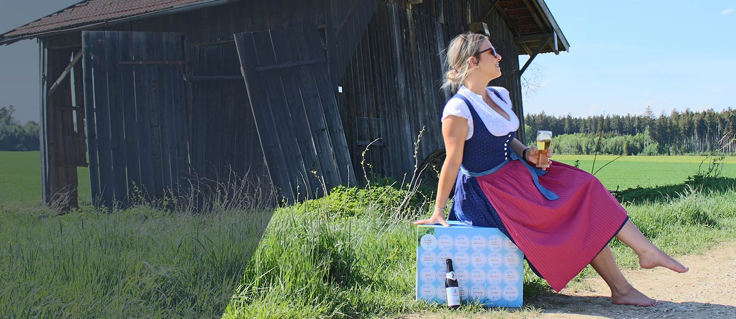 Junge Frau im Dirndl mit Bierglas auf blauer Bierkiste sitzend, vor Scheune und weiter Landschaft, perfekt für bayerische Biergeschenke.