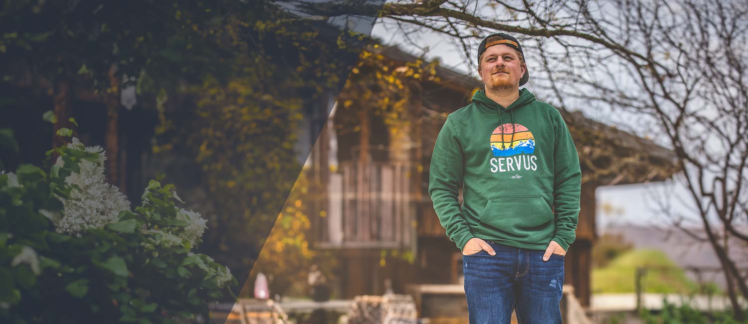 Mann mit grünem bayerischen Hoodie und "Servus" Aufdruck, Jeans und Basecap, im Freien vor rustikalem Haus.