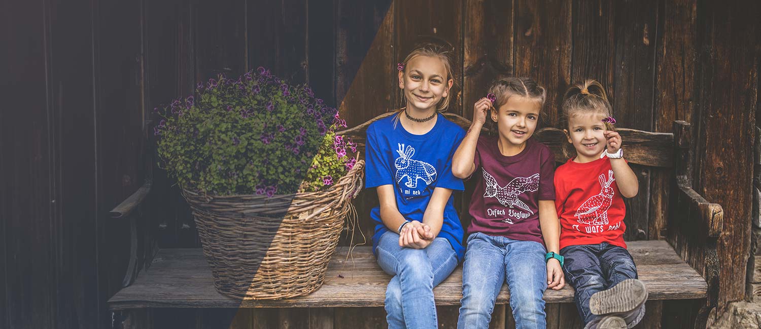 Drei fröhliche Kinder in bayerischen T-Shirts mit Tiermotiven, sitzend auf einer Holzbank.