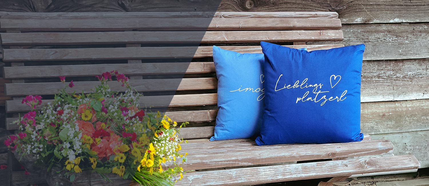 Zwei blaue bayerische Dekokissen mit aufgesticktem Schriftzug „Lieblingsplatzl“ auf einer Holzbank mit Blumenstrauß.