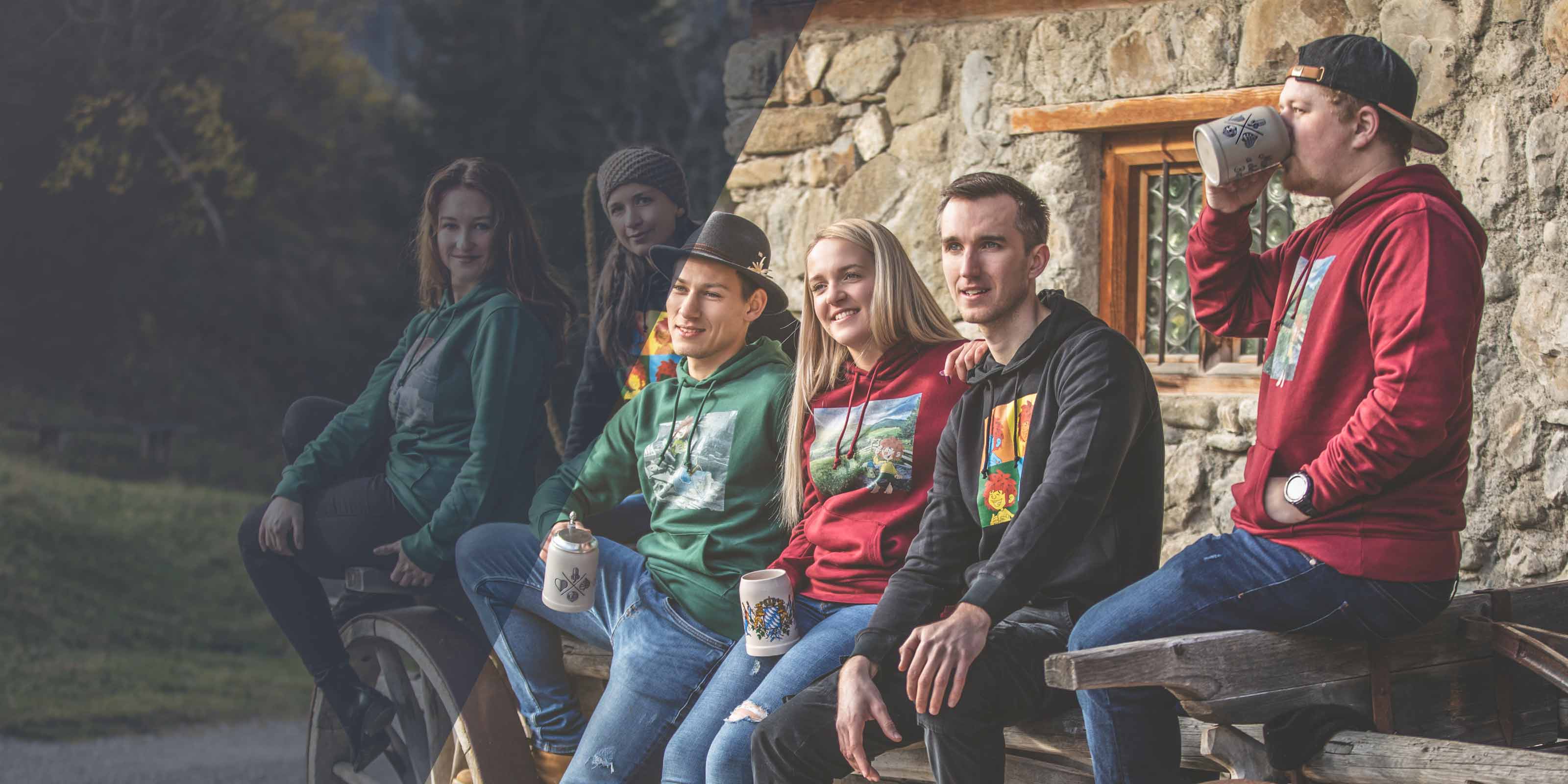 Gruppe von Freunden in Unisex Hoodies mit bayerischen Motiven sitzen auf einer Holzbank vor einem Steinhaus.