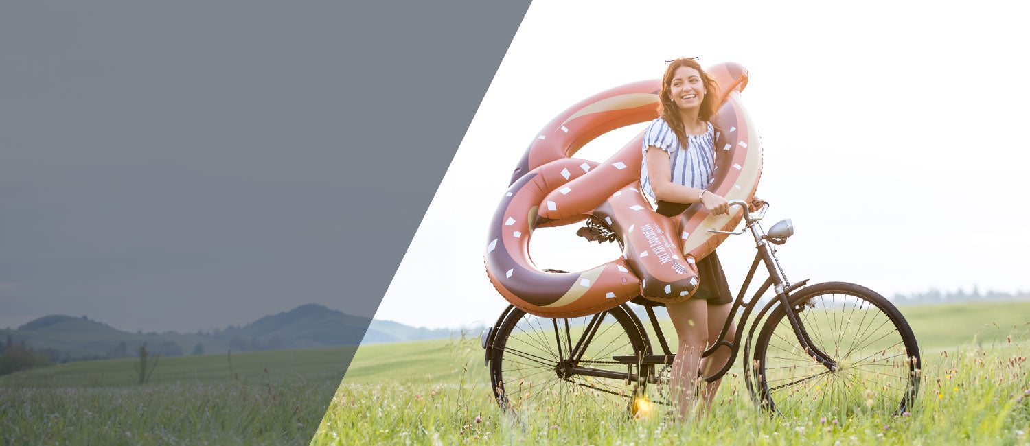 Fröhliche junge Frau mit Brezel Schwimmring auf einem Fahrrad im Grünen, beliebte bayerische Geschenkidee.