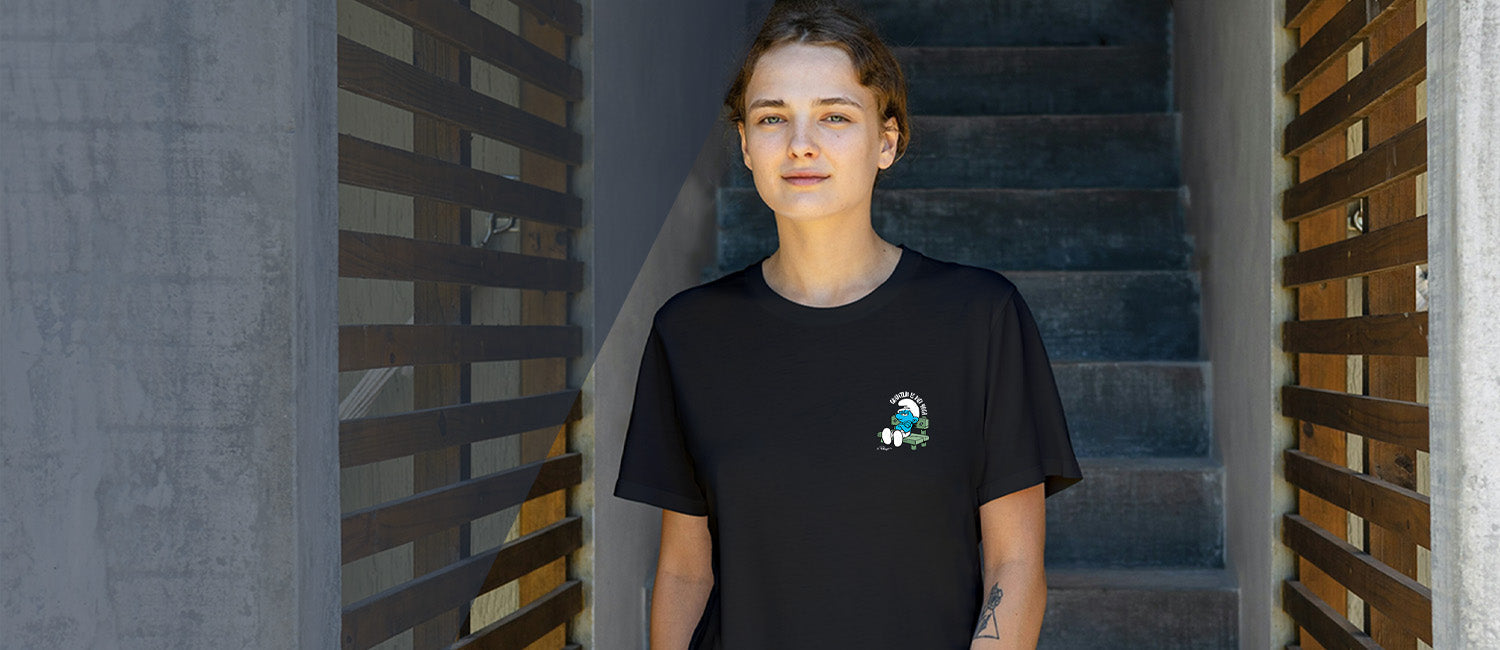 Frau in schwarzem Schlumpf T-Shirt mit kleinem, lächelndem Schlumpf Motiv auf der Brust, sitzend auf einer Bank.