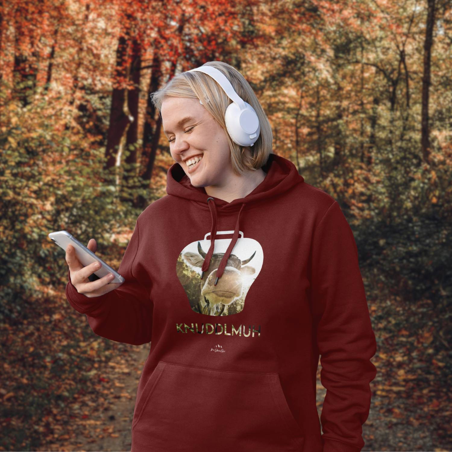 Lächelnde Frau mit weißem Kopfhörer und rotem Hoodie Knuddlmuh mit Kuhmotiv im herbstlichen Wald.