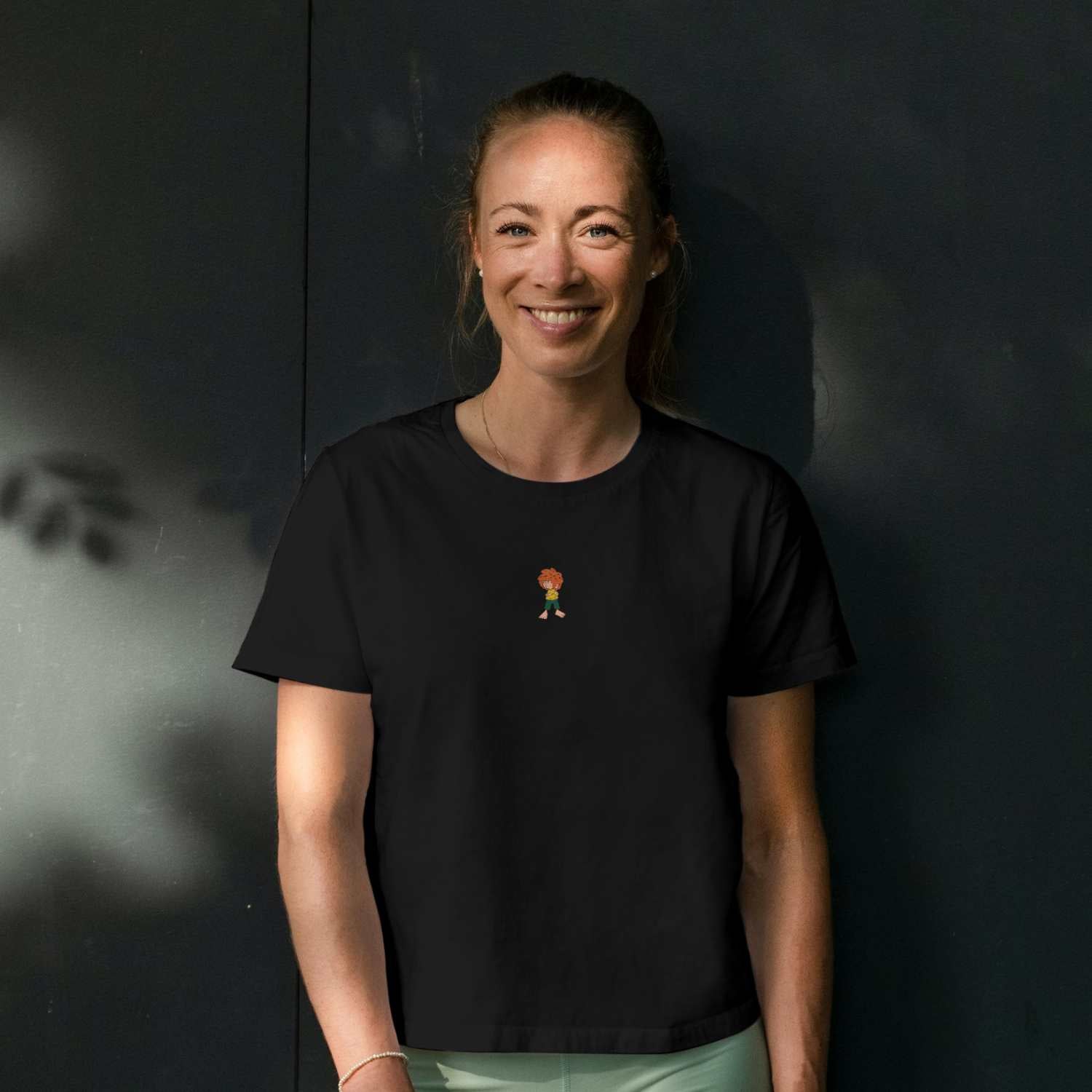 Frau trägt schwarzes Damen T-Shirt mit gewebtem Pumuckl-Motiv auf der Brust, vor dunklem Hintergrund