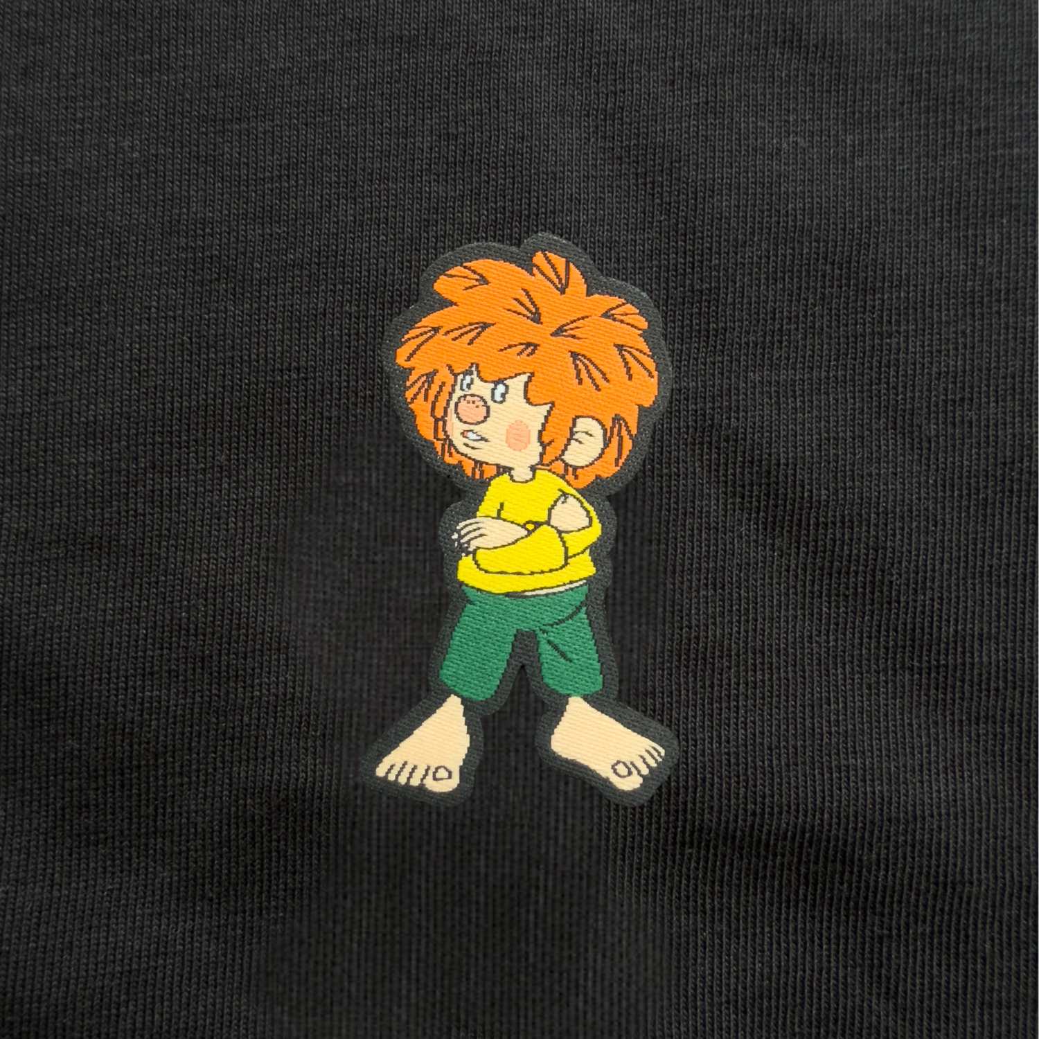 Gewebtes Pumuckl-Label auf schwarzem Damen T-Shirt, zeigt den Kobold mit verschränkten Armen in gelbem Shirt und grüner Hose