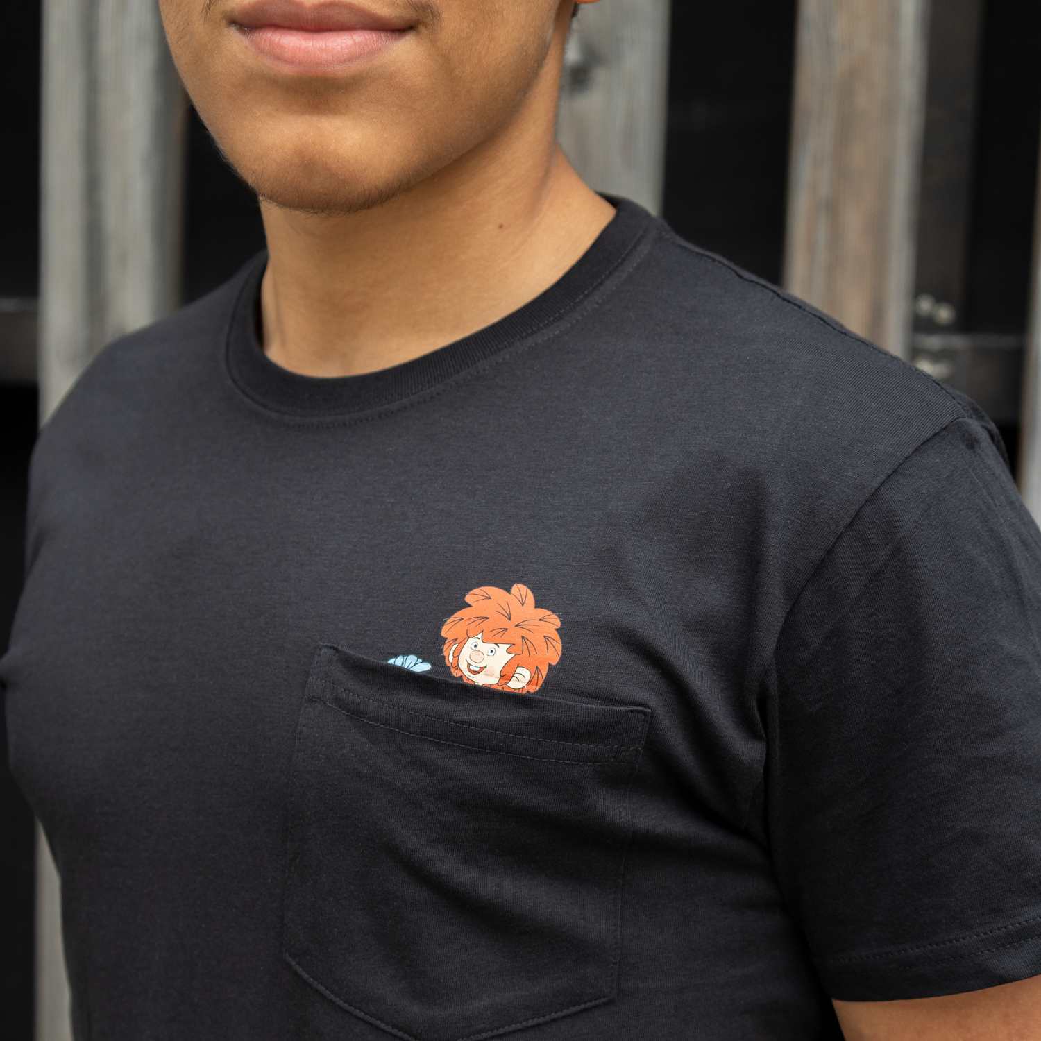 Seitliche Ansicht eines Mannes mit schwarzem T-Shirt, Pumuckl-Motiv blickt aus der Brusttasche hervor.