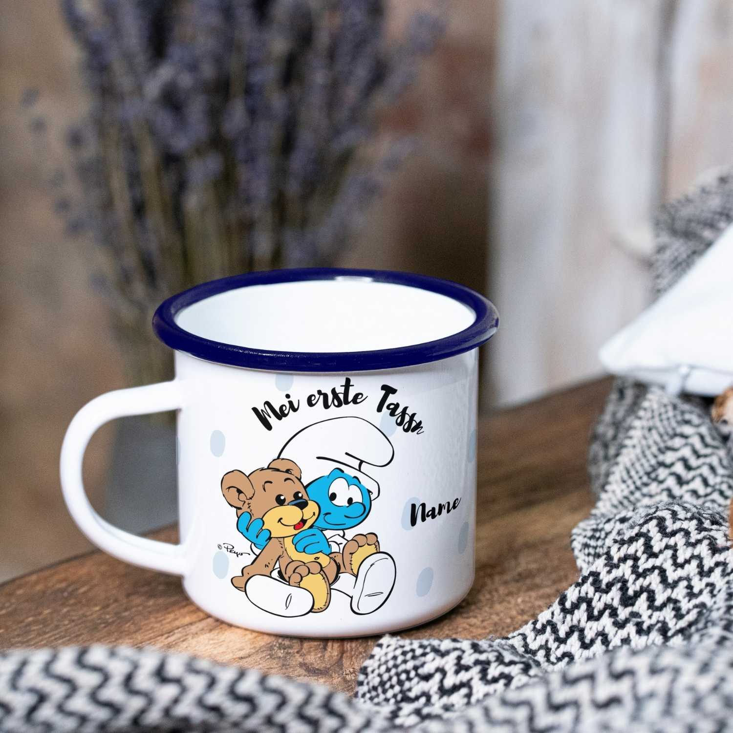 Personalisierbare Emailletasse mit Schlumpf-Motiv, Schriftzug „Mei erste Tassn“ und blauem Rand, Füllmenge 330 ml.