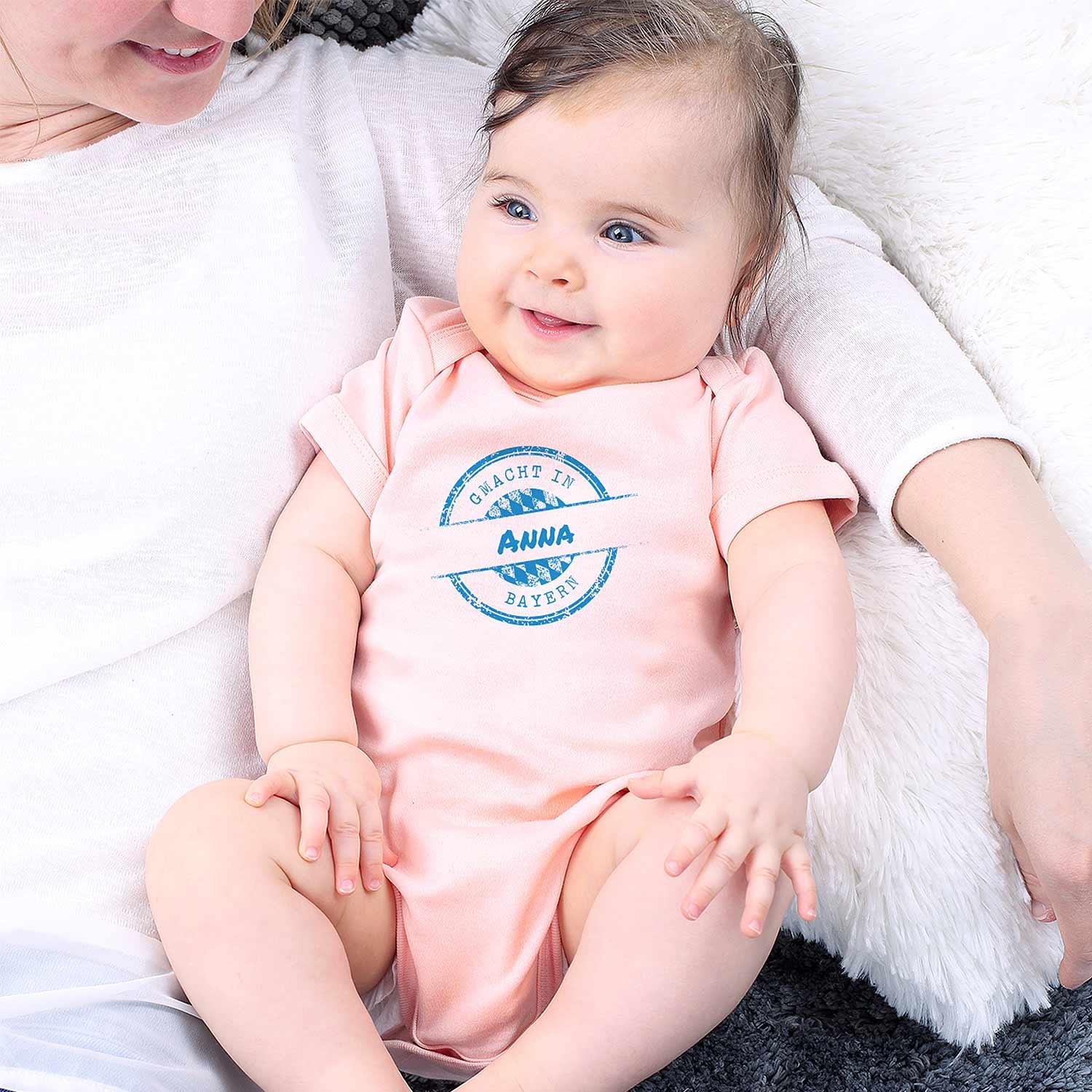 Rosa Baby-Body mit weißem Aufdruck „gmacht in Bayern“ und personalisierbarem Wunschname, kurze Ärmel, Druckknöpfe unten, Baumwolle.