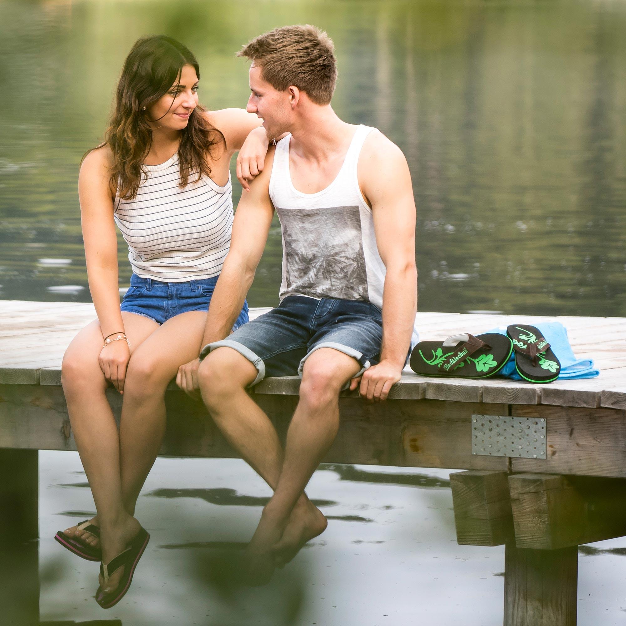 Glückliches junges Paar sitzt entspannt auf einem Holzsteg am Seeufer im Sommerurlaub.