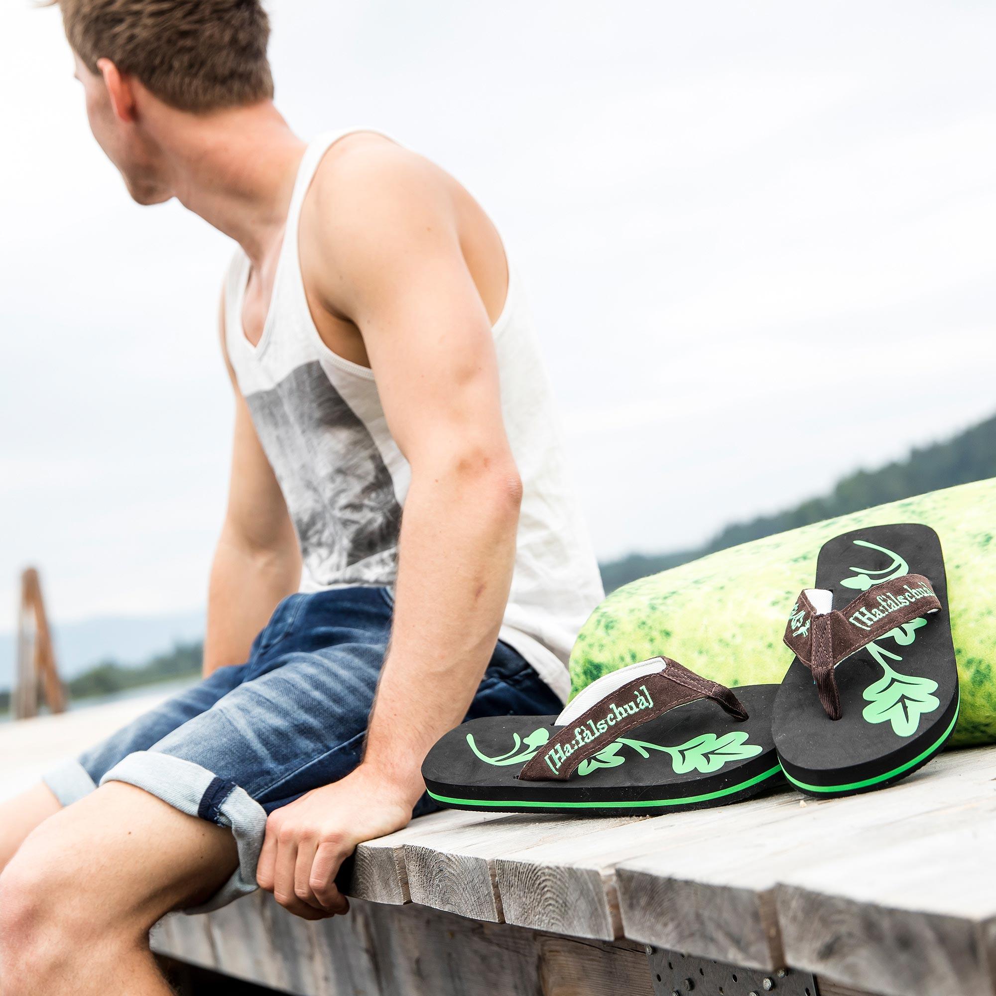 Sommerliches Paar Flip-Flops in Schwarz-Grün mit braunen Riemen und Eichenblattmotiv auf Holzsteg.