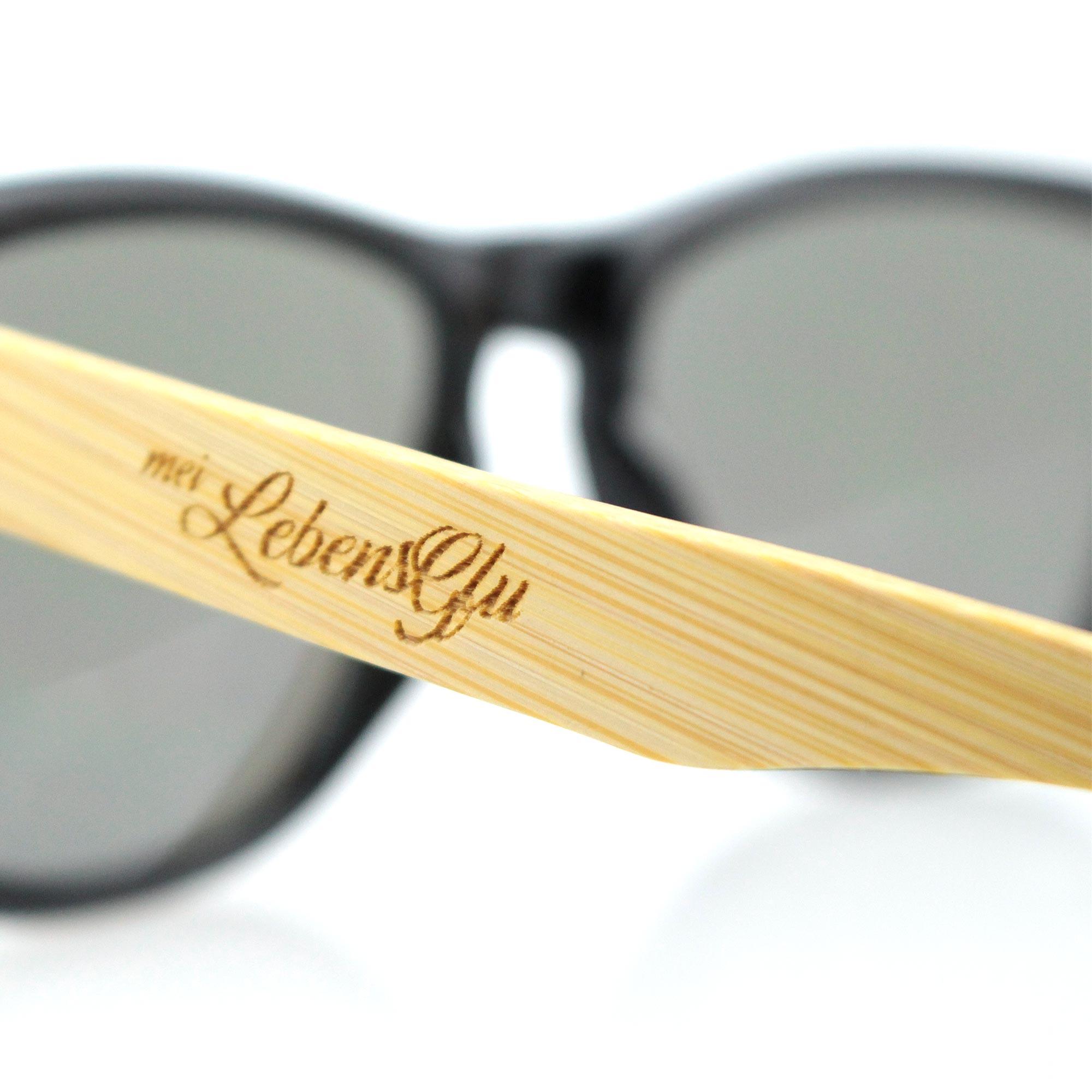 Sonnenbrille mit schwarzem Kunststoffrahmen, Bügeln aus Bambus und Gravur „Gäh mia aus da Sunn!“ auf dem Holz.