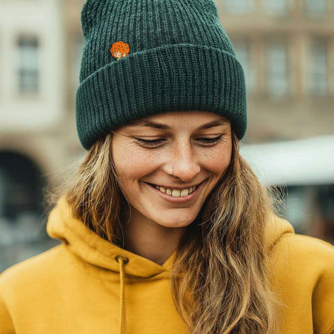 Lächelnde Frau trägt die Pumuckl Beanie aus Baumwolle in Dunkelgrün mit gewebtem Pumuckl-Label und gelbem Hoodie