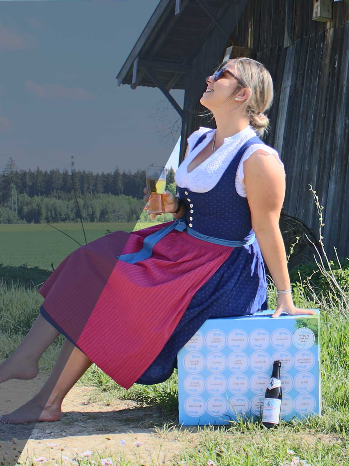 Traditionell gekleidete Frau im Dirndl mit einem Radler an einem Holzlager vor Waldkulisse.