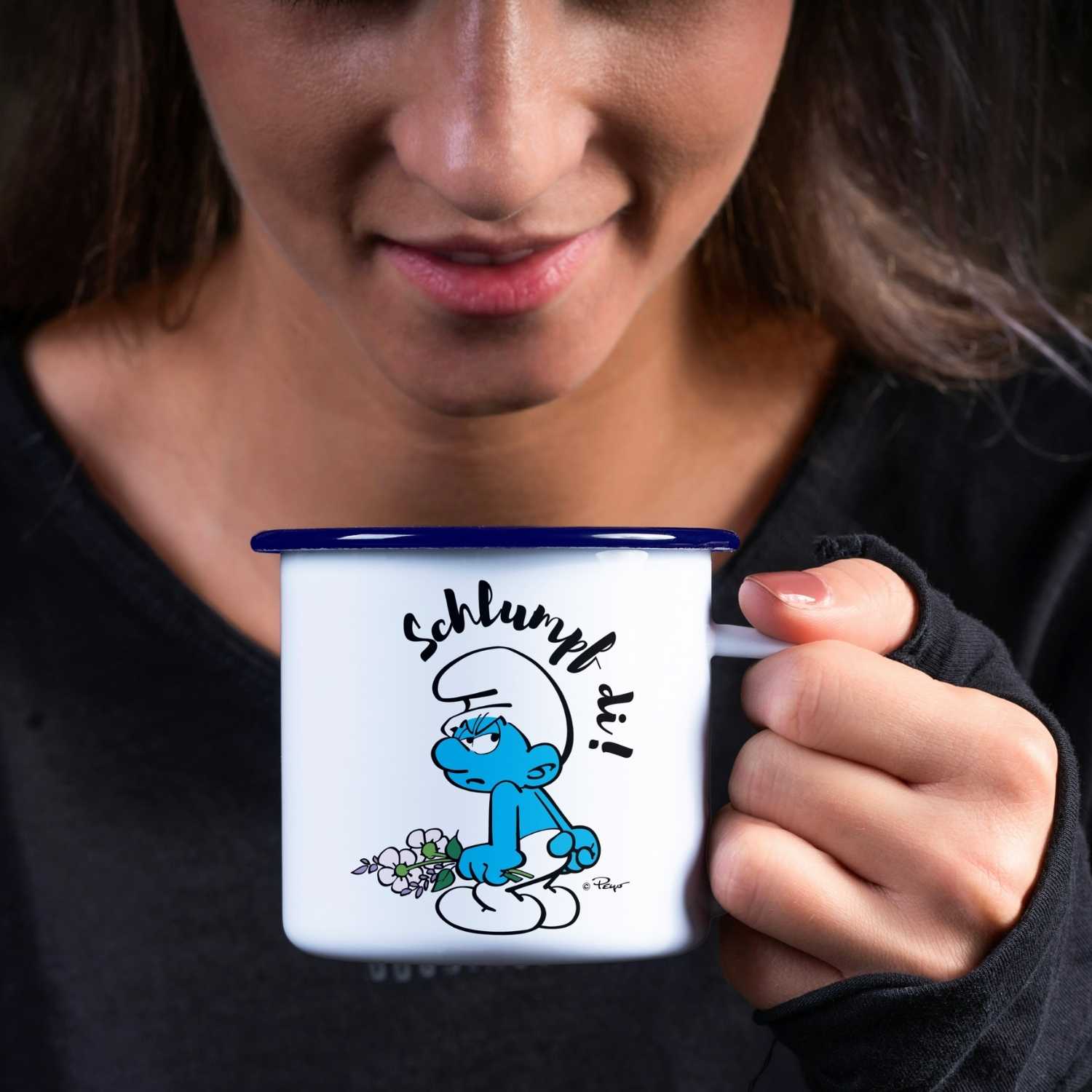 Frau hält eine ®Die Schlümpfe Emailletasse Schlumpf di! mit miesepetrigem Schlumpf und Blumenmotiv fest.