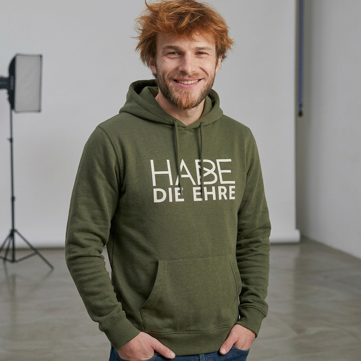 Lächelnder Mann mit rotem Haar trägt olivgrünen Hoodie Habe die Ehre Aufdruck und Kapuze.