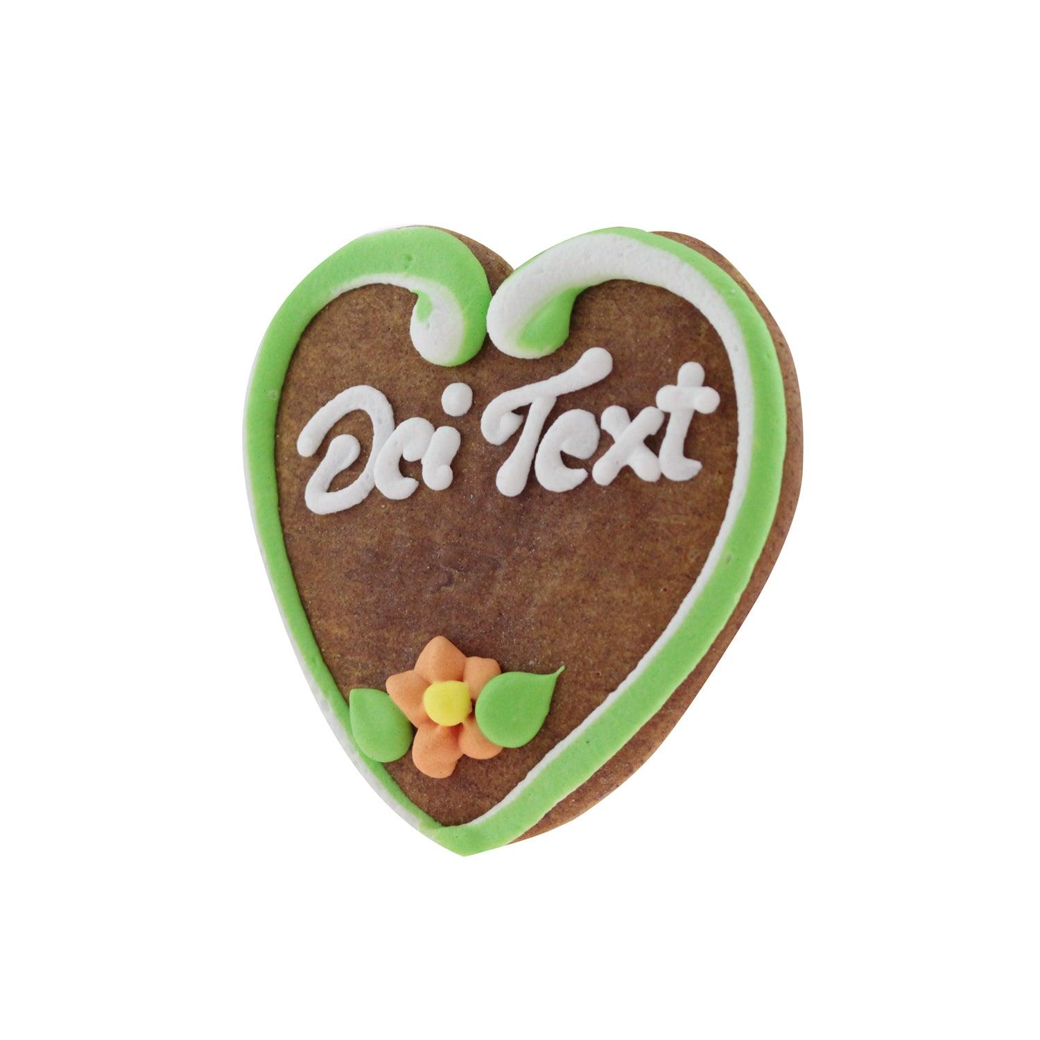 Herzförmiges Lebkuchenherz mit weißem Randdekor, rosa Zuckerschrift und zwei kleinen Blumen aus Zuckerguss, ca. 10 x 10 cm.