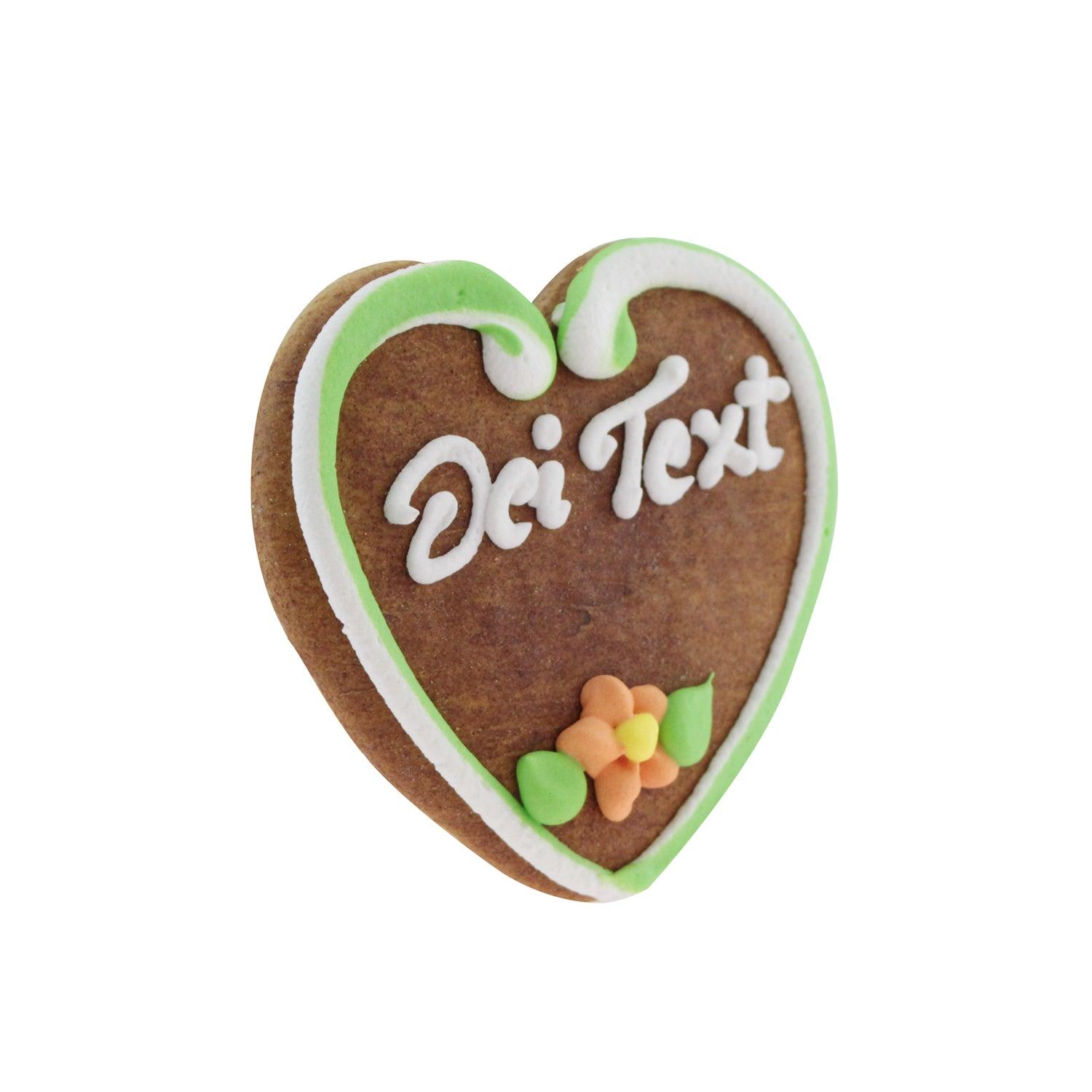 Braunes Lebkuchenherz mit weiß-rotem Zuckerrand, rosa Blumen und rotem Schriftzug auf glattem Zuckerguss; ca. 10 × 10 cm.