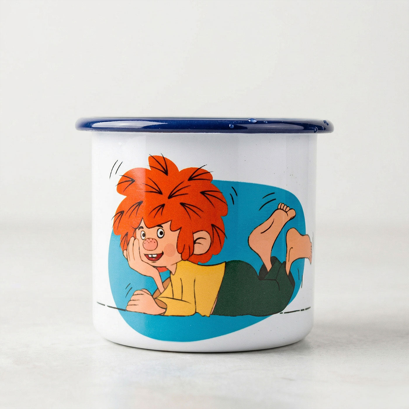 Weiße Emaille-Tasse mit blauem Rand und buntem Pumuckl-Motiv, der schaumige Blicke wirft.