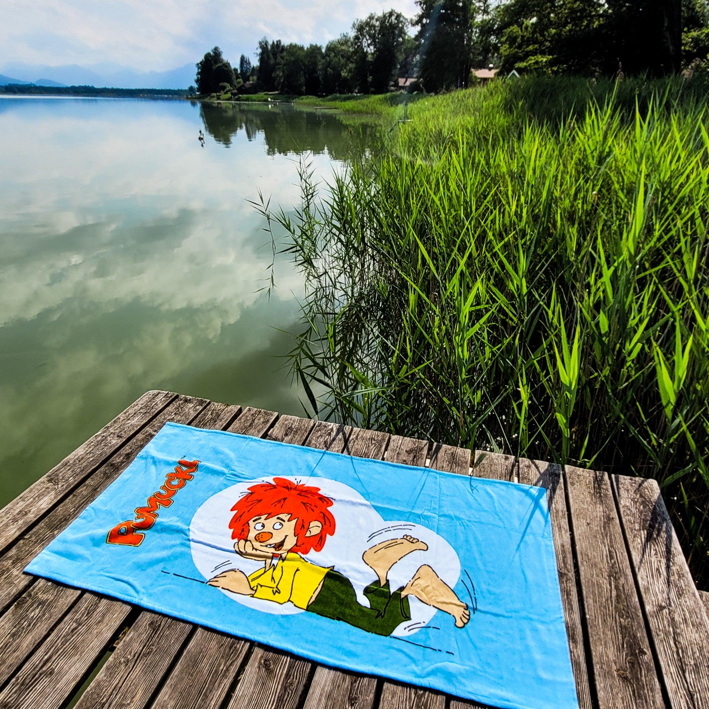 Pumuckl Badetuch XXL auf hölzernem Steg am See mit Schilf und Bäumen, perfekte Badeutensilien für Kinder.