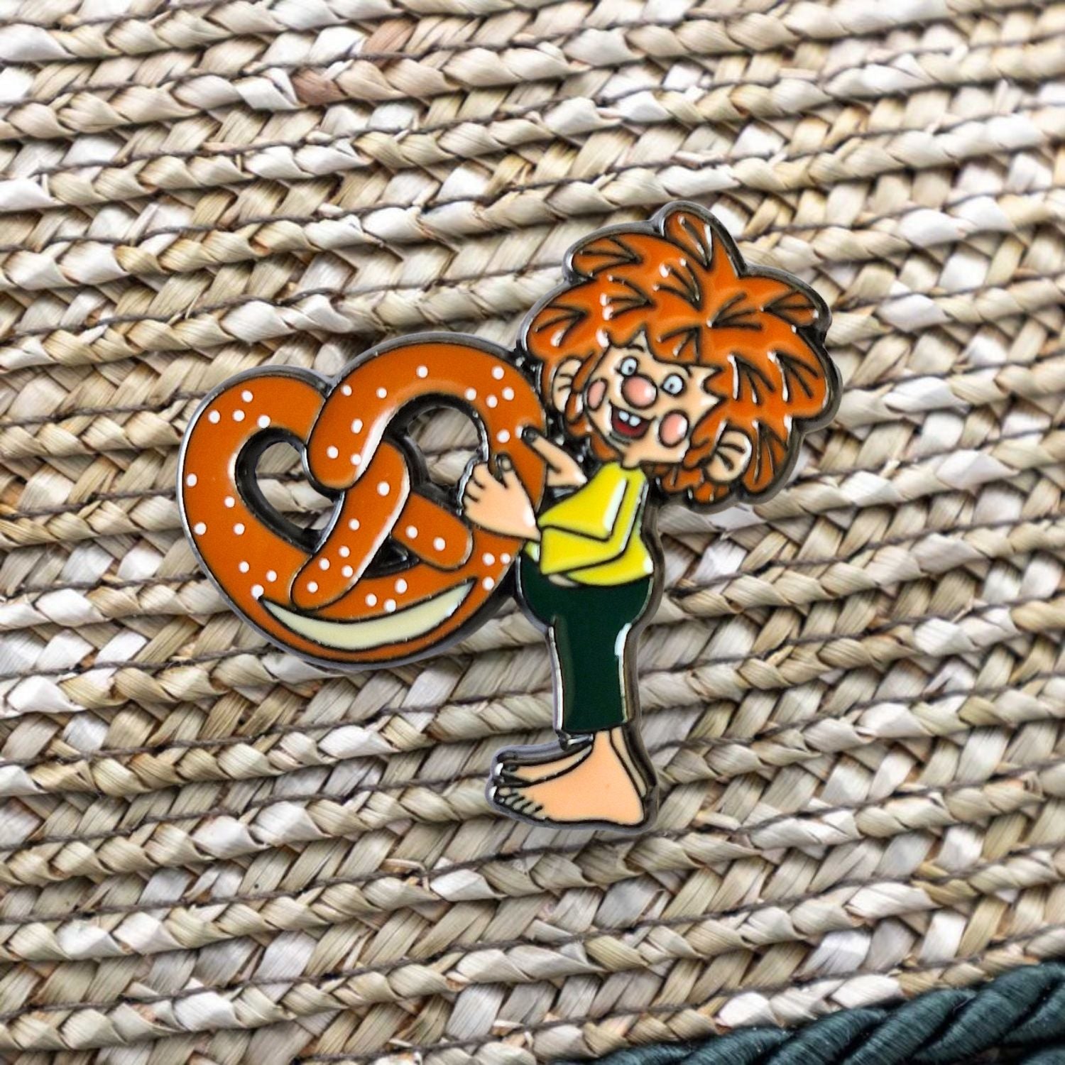 Lebhafter Pumuckl Pin mit roter Haarpracht, der eine große gesalzene Brezel umarmt, platziert auf einem geflochtenen Untergrund.