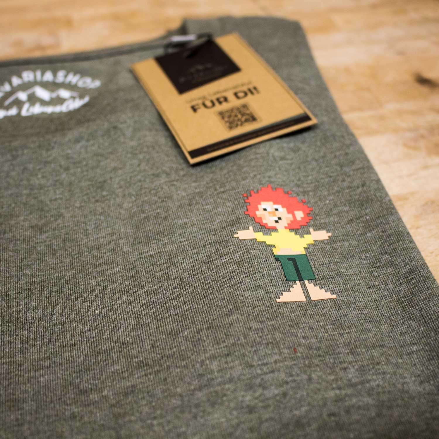 Grünes Baumwoll-T-Shirt Pixmuckl mit farbigem Pixel Pumuckl-Motiv und Etikett auf Holzuntergrund.