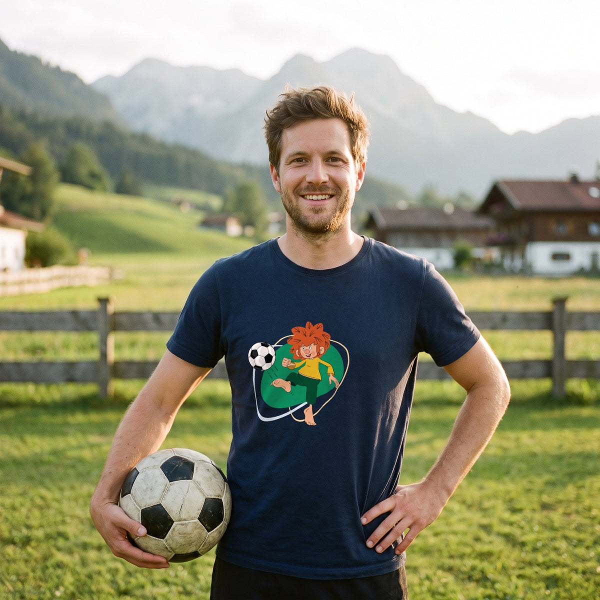 Mann im dunkelblauen Pumuckl T-Shirt Herren „Fußball Edition“ mit Fußball unterm Arm auf einer Wiese vor bayerischen Bergen – Pumuckl kickt barfuß den Ball