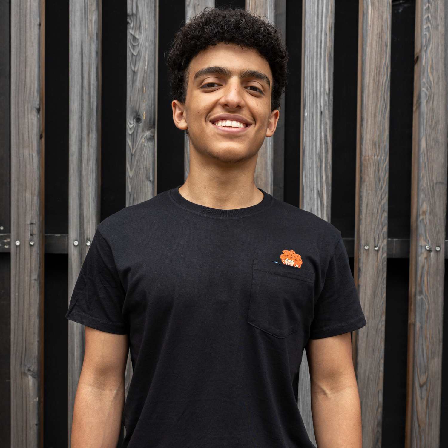 Lächelnder Mann trägt schwarzes Pumuckl T-Shirt Schabernack mit Brusttasche und rotem Kobold-Emblem, vor Holzwand.