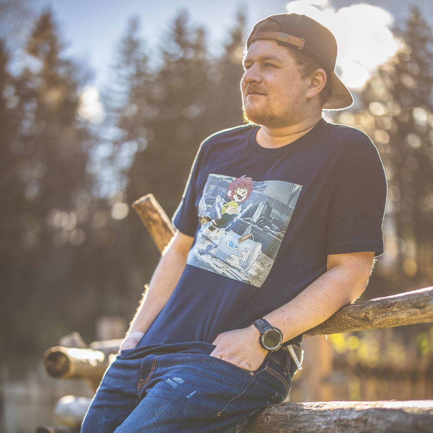 Mann trägt dunkelblaues Pumuckl Herren T-Shirt Werkstatt mit Motiv, Jeans und Cap im Freien.