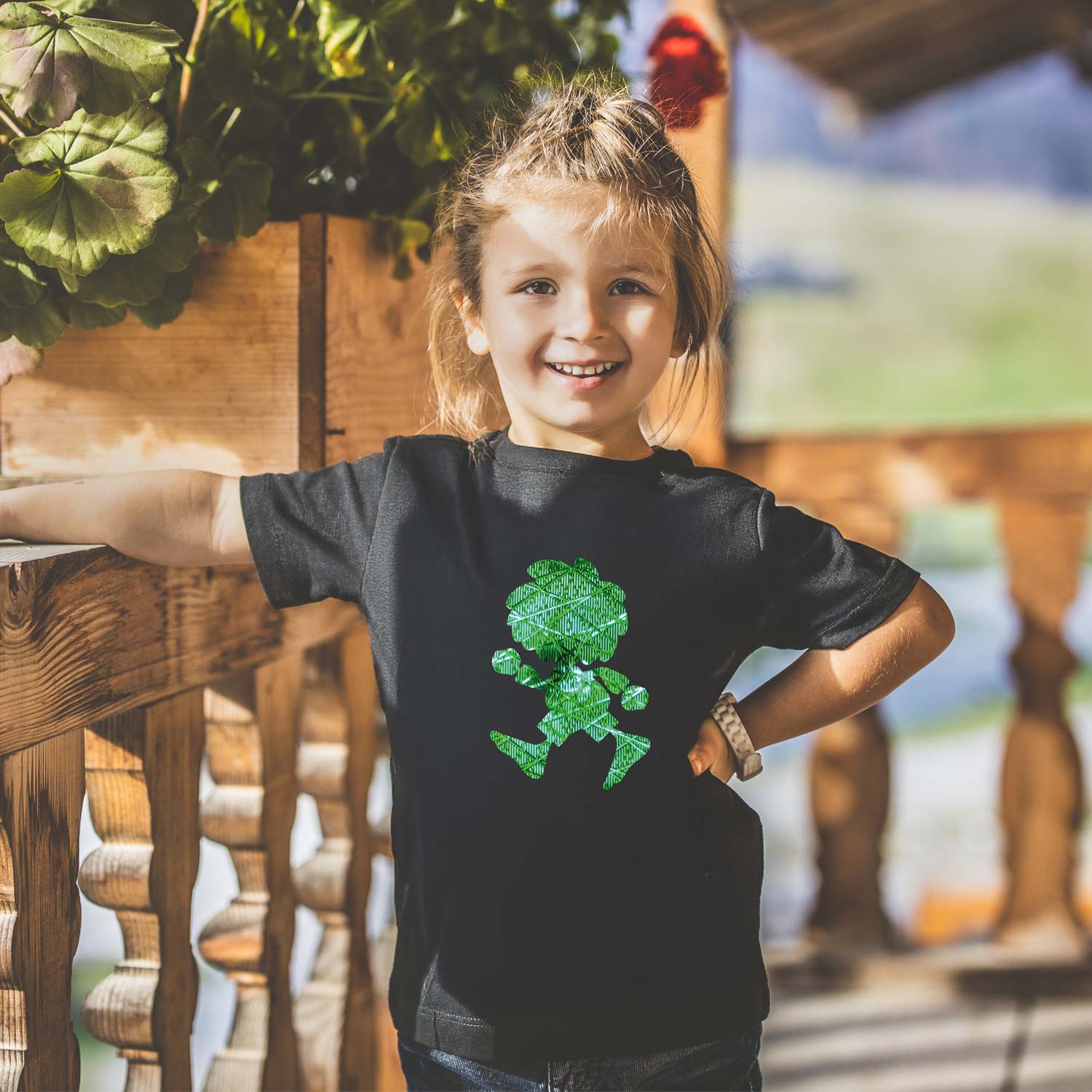 Lachendes kleines Mädchen in schwarzem Pumuckl Kinder T-Shirt Ampel-Edition mit Holzgeländer und Pflanzen im Hintergrund