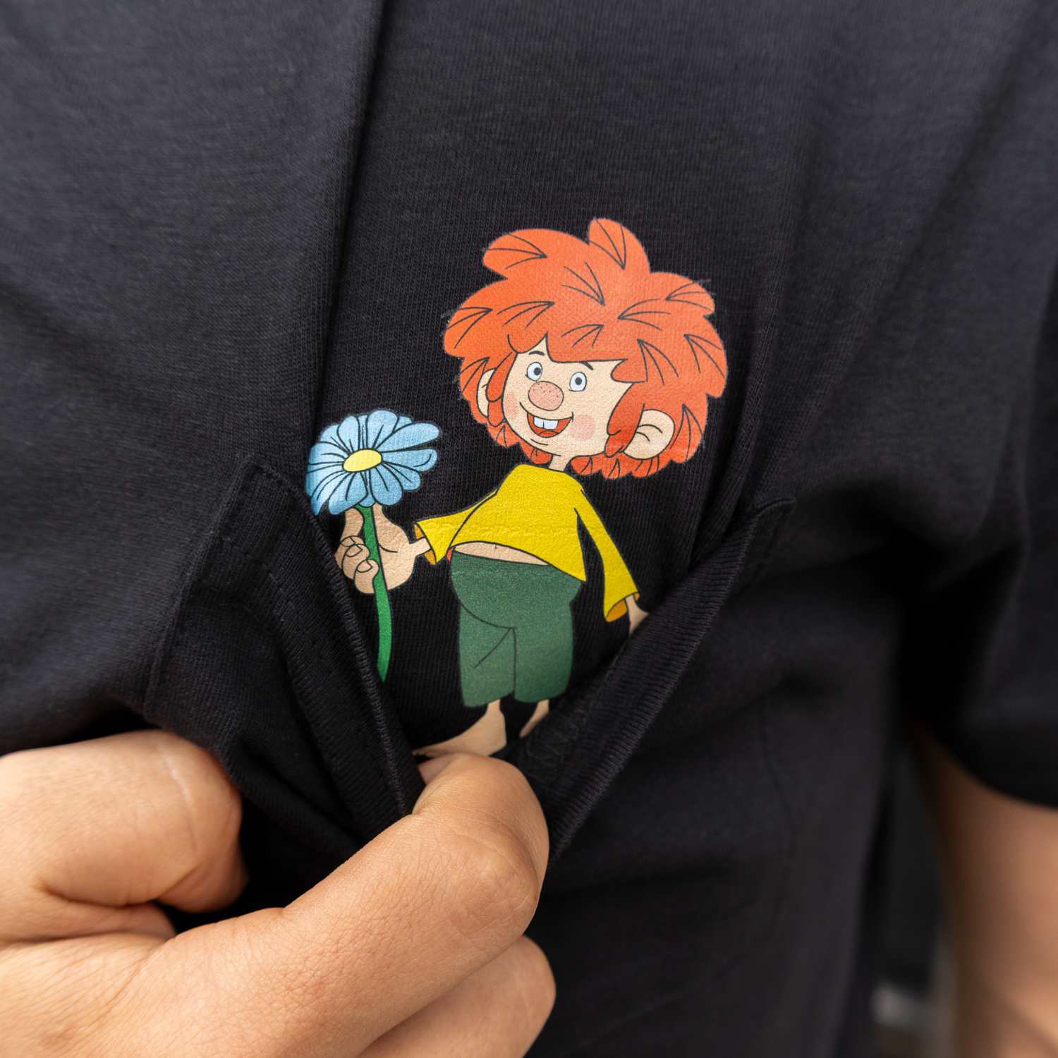 Nahaufnahme des Pumuckl Motivs auf einem schwarzen T-Shirt, der Kobold steckt in einer Tasche und hält eine blaue Blume hoch.