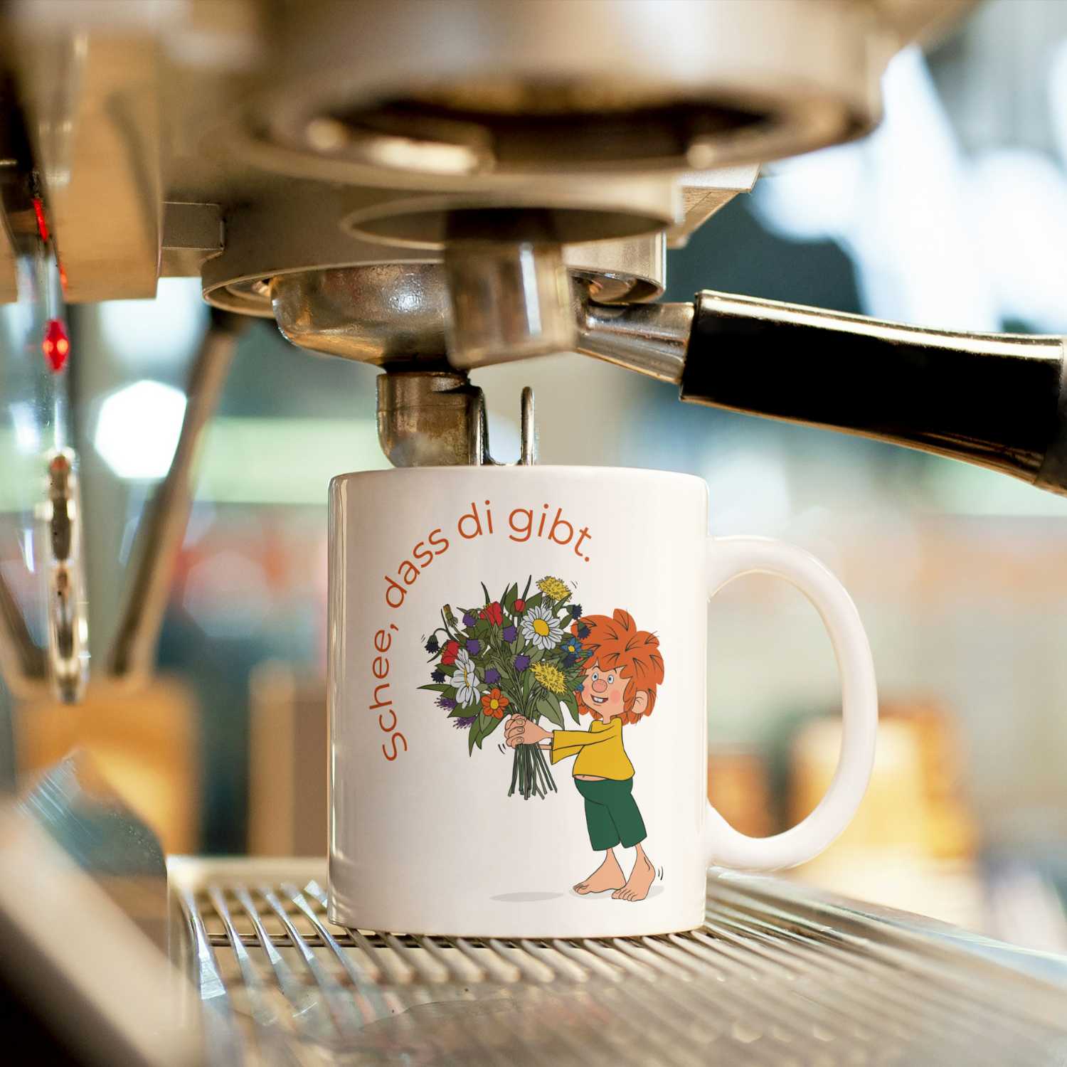 Weiße ®Pumuckl Tasse „Schee, dass di gibt.“ auf Espressomaschine, mit Blumenstrauß-Motiv.
