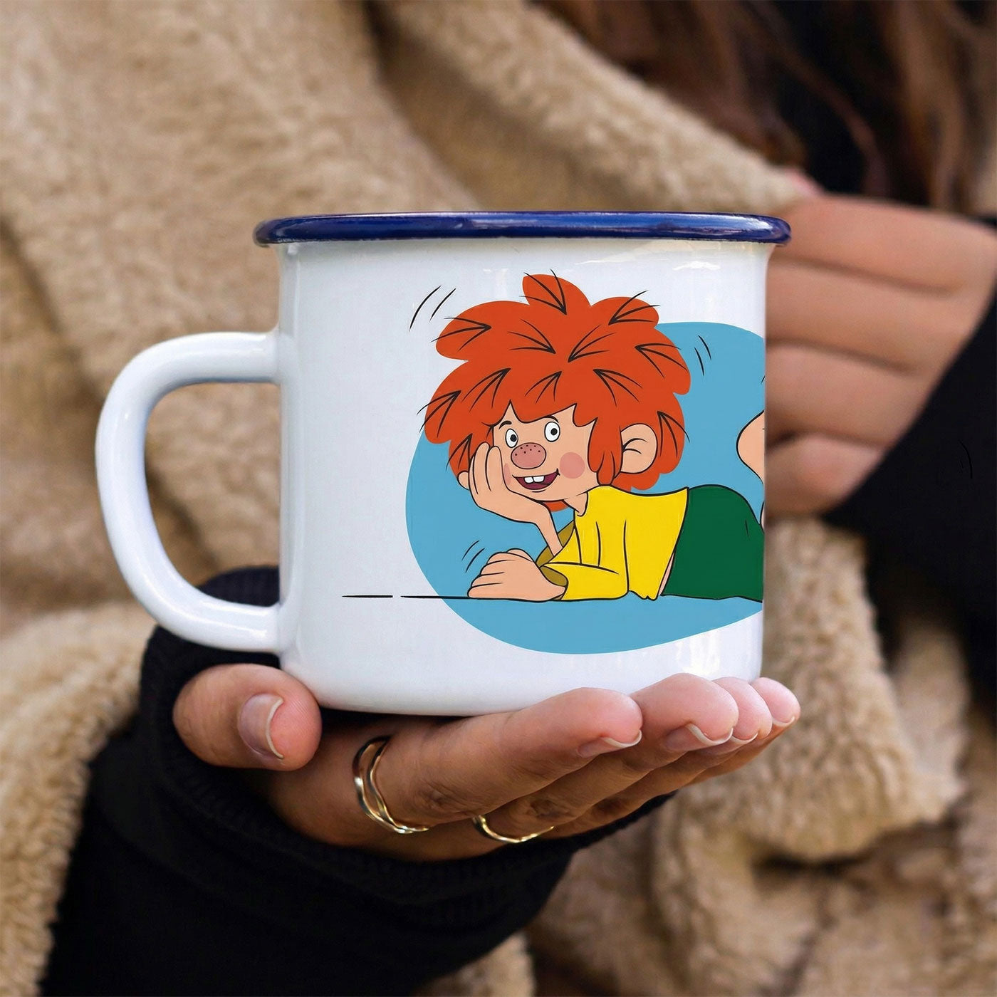 Hände halten eine weiße Emaille-Tasse mit blauem Rand und Pumuckl-Motiv, aufgeregt liegend.