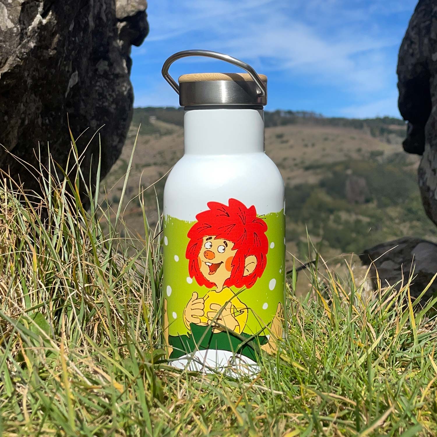Pumuckl Thermo Trinkflasche aus Edelstahl mit rotem Kobold-Motiv, Holzgriff, in Graslandschaft mit Felshintergrund.