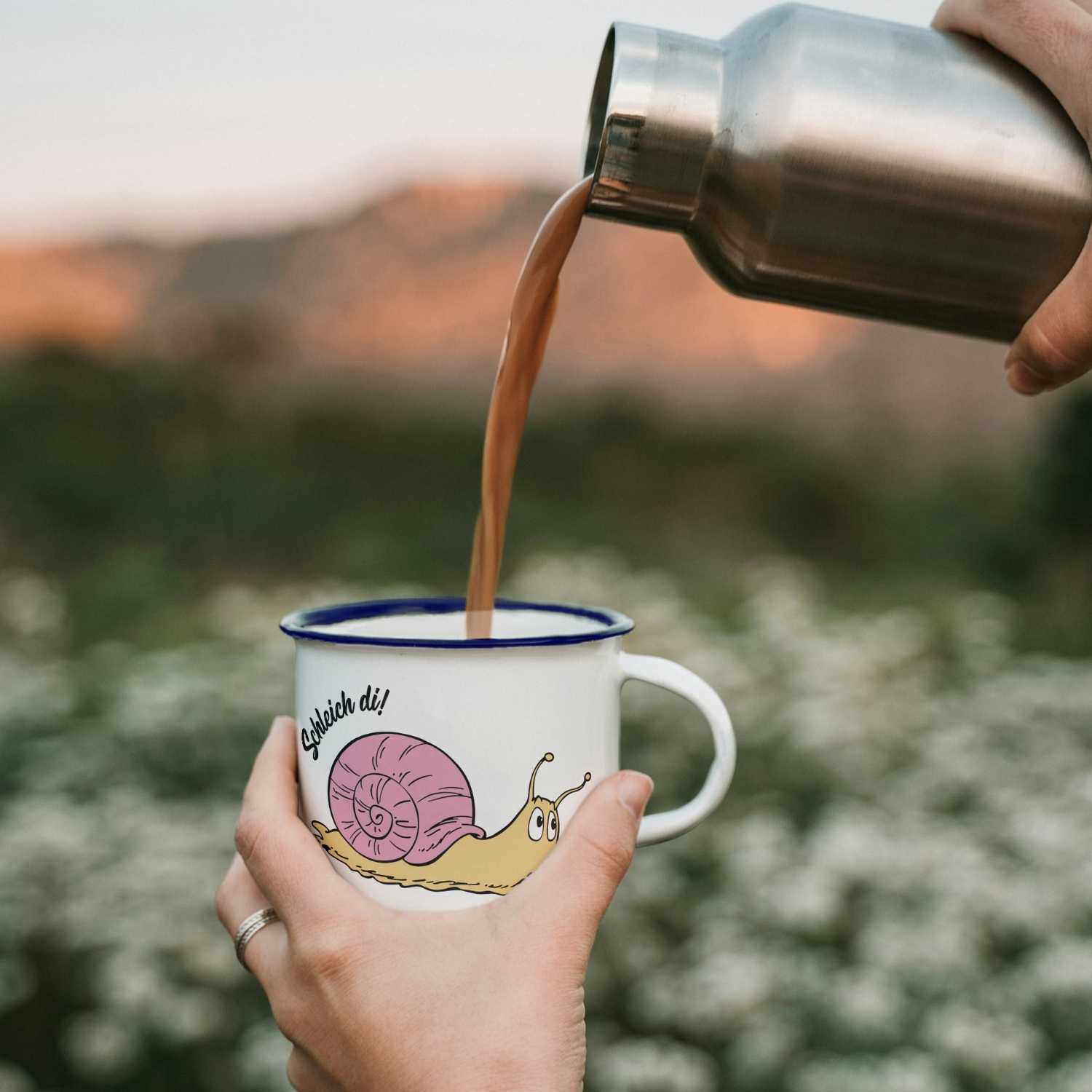 ®Die Schlümpfe Emailletasse "Schleich di" mit Schneckenmotiv, von Hand mit heißem Getränk gefüllt vor Bergkulisse.