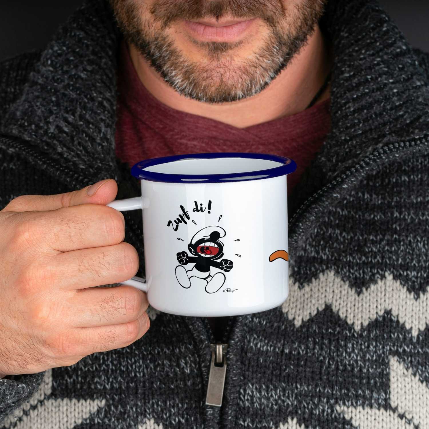 Mann hält ®Die Schlümpfe Emailletasse "Zupf di!" mit Comic-Motiv eines lachenden Schlumpfes.