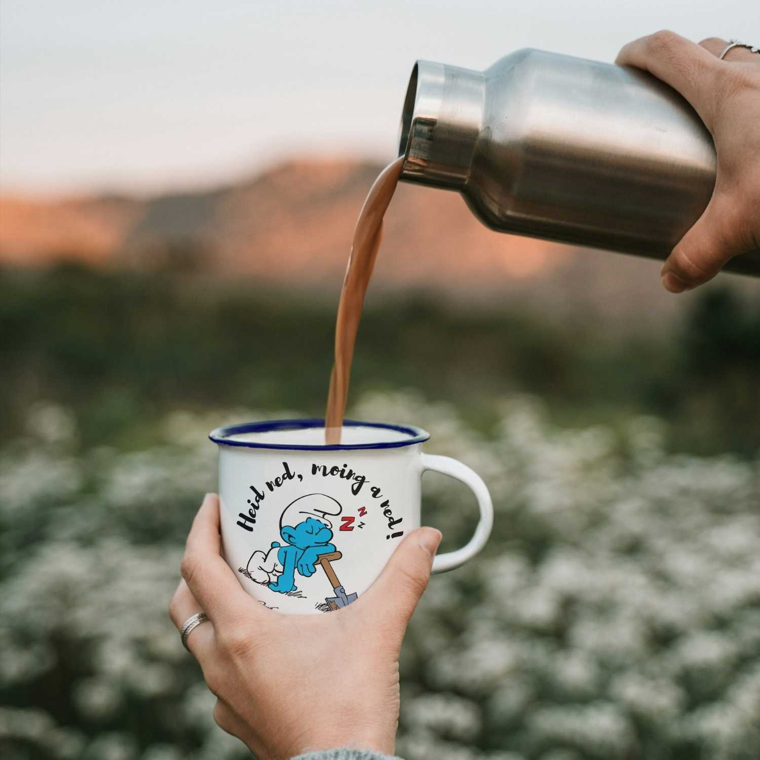 Hand hält Die Schlümpfe Emailletasse Heid ned moing a ned, braunes Getränk wird aus Thermosflasche gegossen.