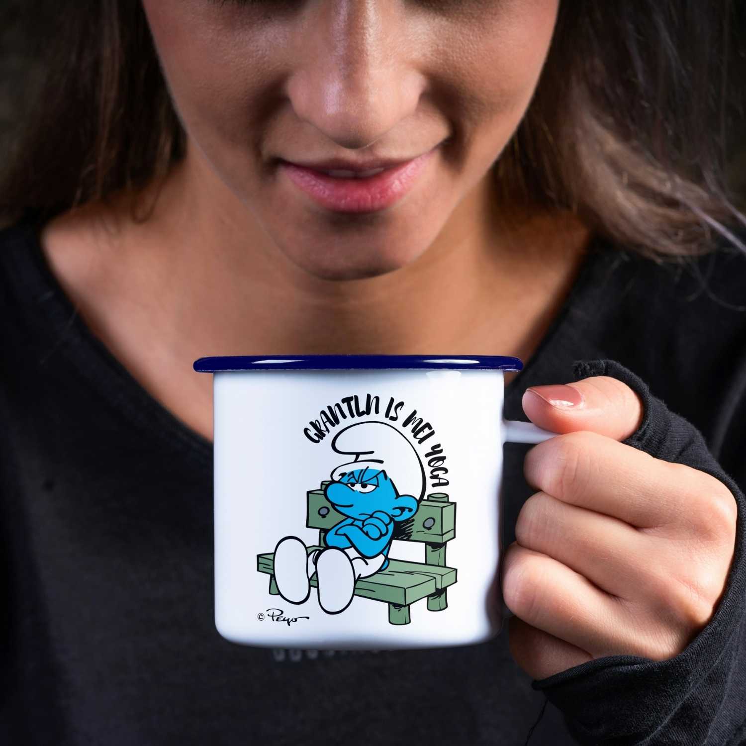 Dunkelhaarige Frau hält emaille Schlümpfe Tasse "Grantln is mei Yoga" mit mürrischem Schlumpf Motiv.