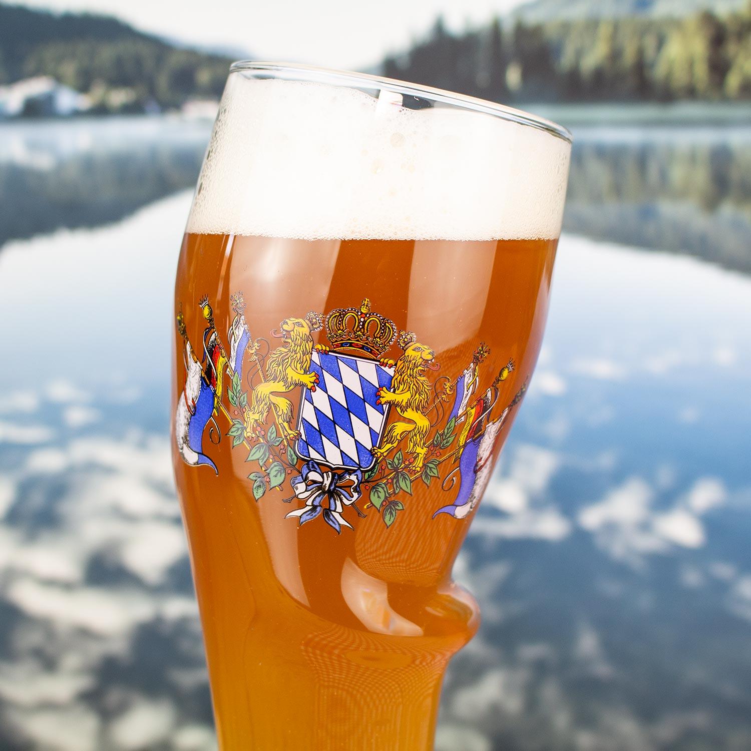 Weißbierglas mit bayerischem Wappen, gefüllt mit goldgelbem Weizenbier und Schaumkrone, vor See-Panorama