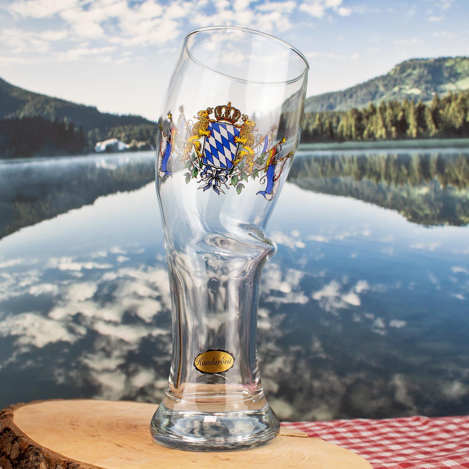 Klares Bierglas mit bayerischem Staatswappen, auf einer Holzscheibe vor Gebirgssee-Kulisse.