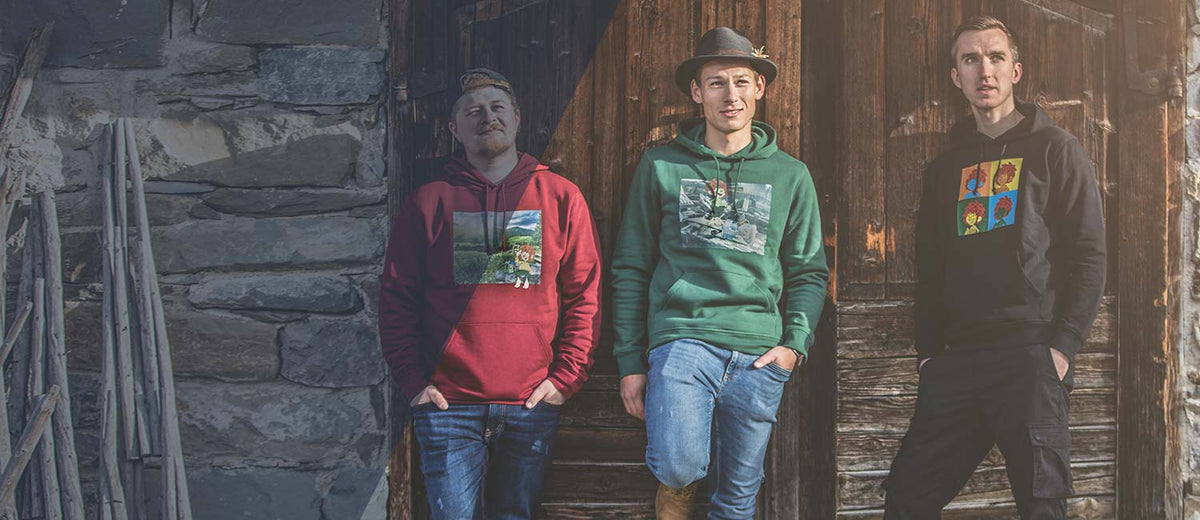 Drei Männer präsentieren Pumuckl Hoodies in Rot, Grün und Schwarz mit farbenfrohen Motiven vor rustikalen Holztüren.