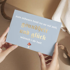 Hände halten die hellblaue Glückwunschkarte "Zum Oidwern...", verziert mit goldener Schrift und einem Fliegenpilz-Motiv.