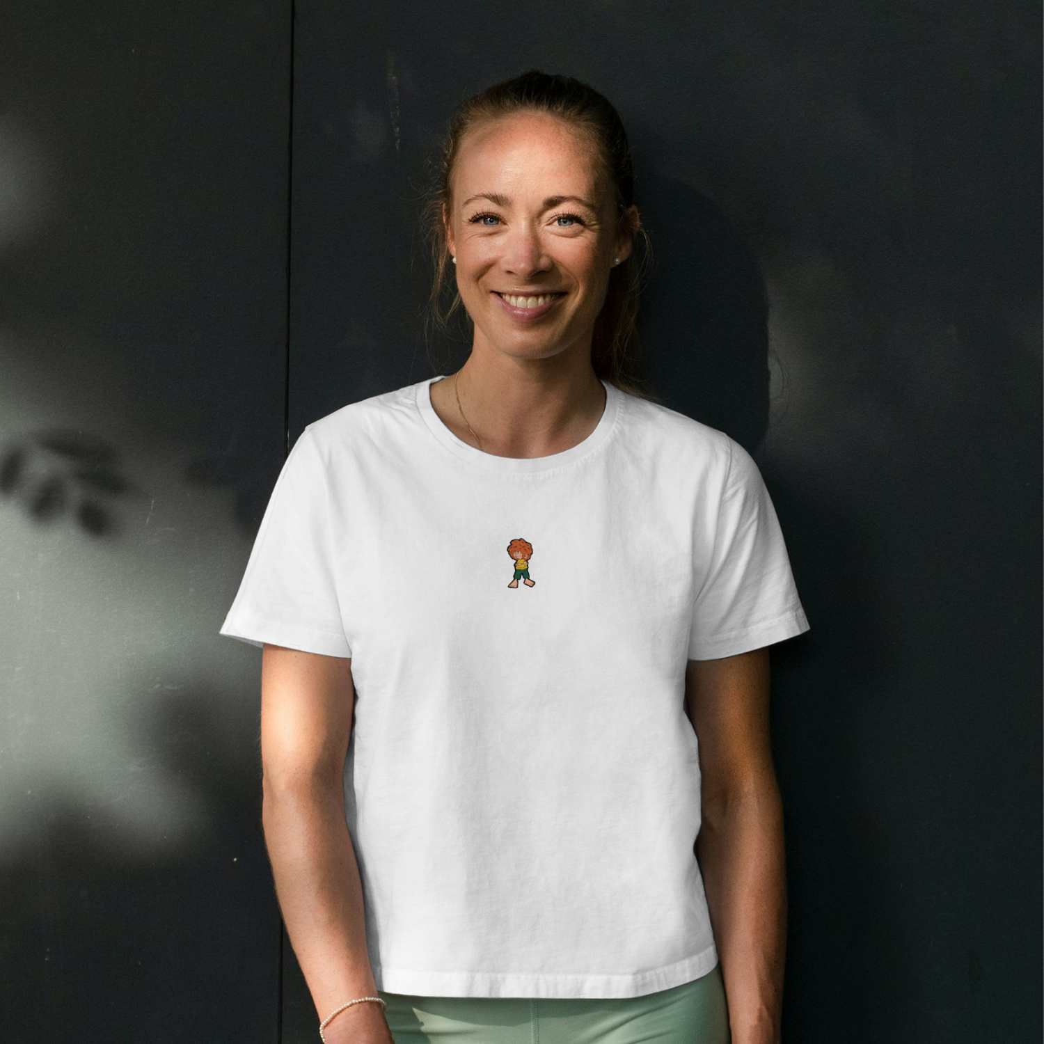 Frau trägt weißes Damen T-Shirt mit gewebtem Pumuckl-Motiv auf der Brust, vor dunklem Hintergrund