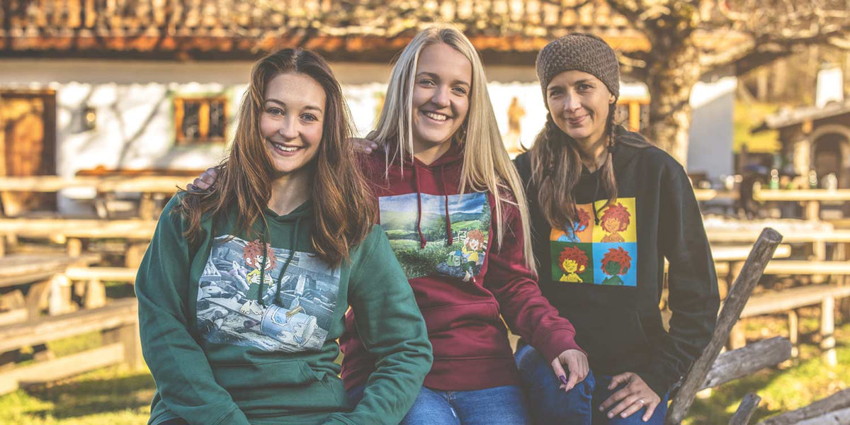 Drei lächelnde Frauen in verschiedenen Hoodies mit Comic-Motiven und bunten Designs im Freien.