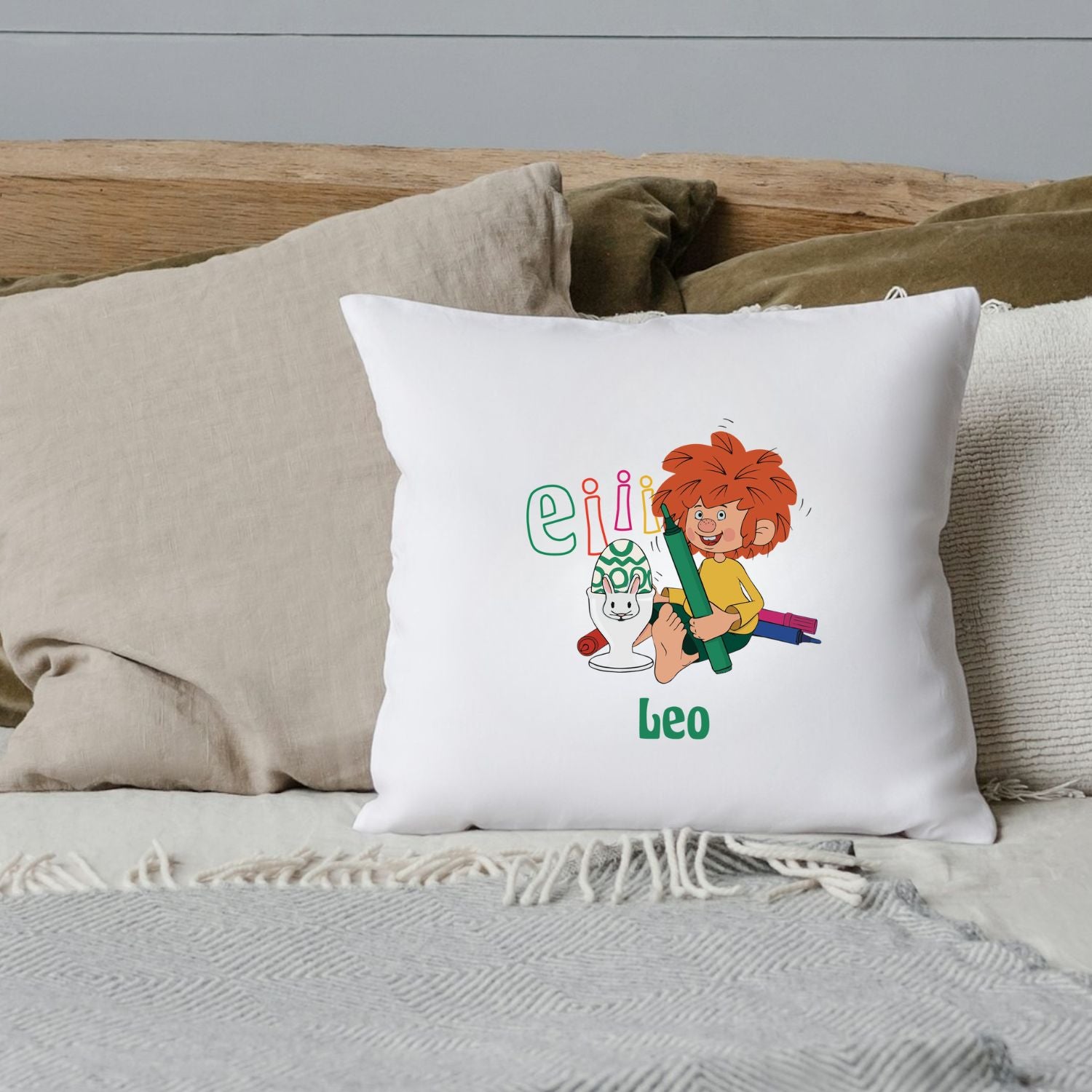 Weißes Pumuckl Kuschelkissen Eiii Ostern mit buntem Pumuckl-Motiv und Name Leo auf einem Bett.