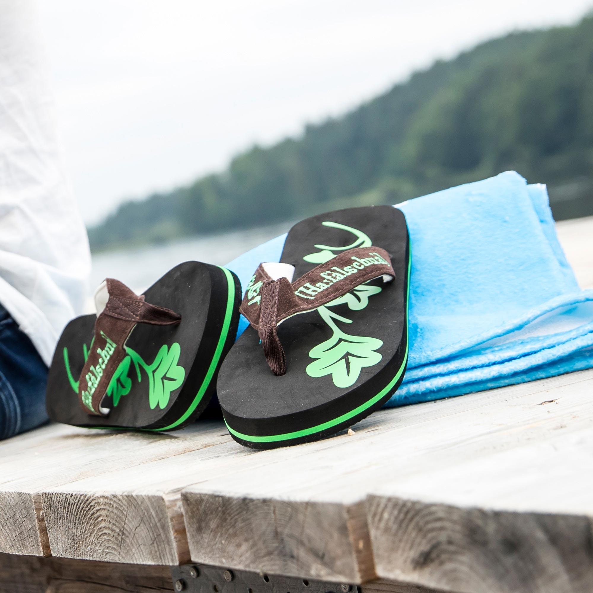 Schwarze Flip-Flops mit grüner Sole und braunen Riemen, daneben ein blaues Handtuch auf Holzsteg am See