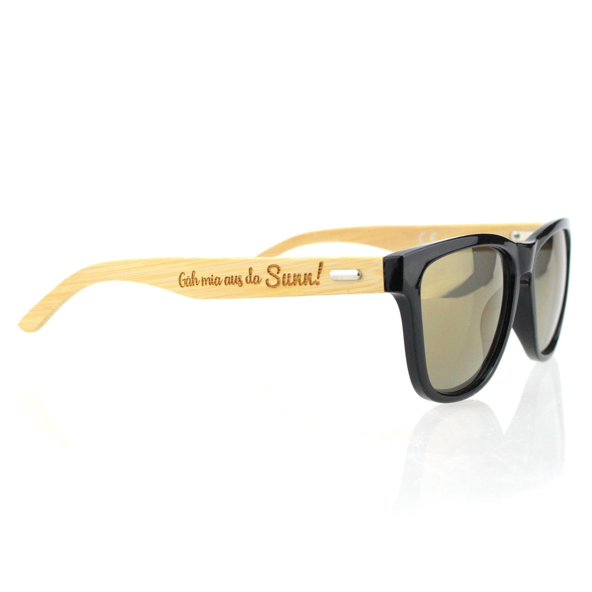 Sonnenbrille mit schwarzem Kunststoffrahmen, Bambus-Holzbügeln und Gravur „Gäh mia aus da Sunn!“ auf dem Bügel.