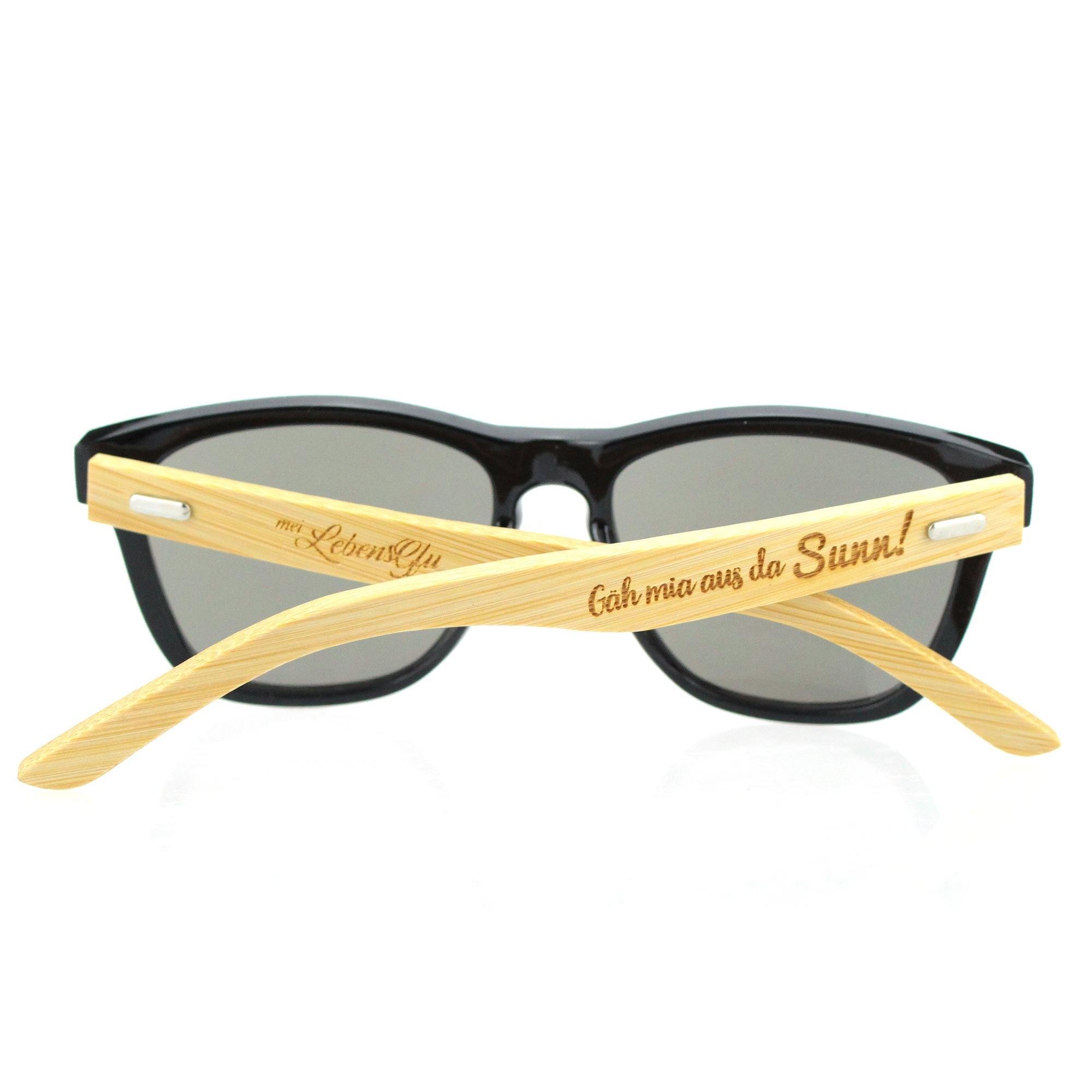 Sonnenbrille mit schwarzem Kunststoffrahmen, Bambusbügeln und Gravur „Gäh mia aus da Sunn!“ auf dem Bügel.