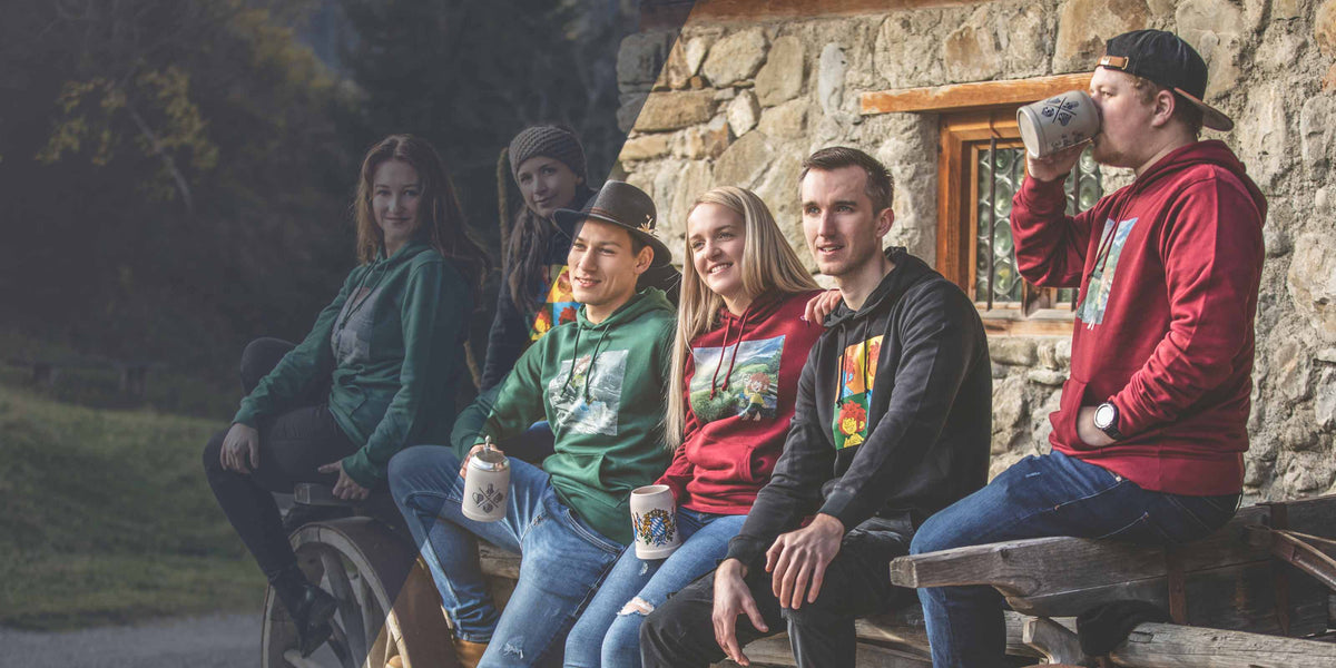 Gruppe junger Erwachsener in farbigen Hoodies mit unterschiedlichen Designs, einige halten Bierkrüge, sitzend vor rustikalem Steinhaus.
